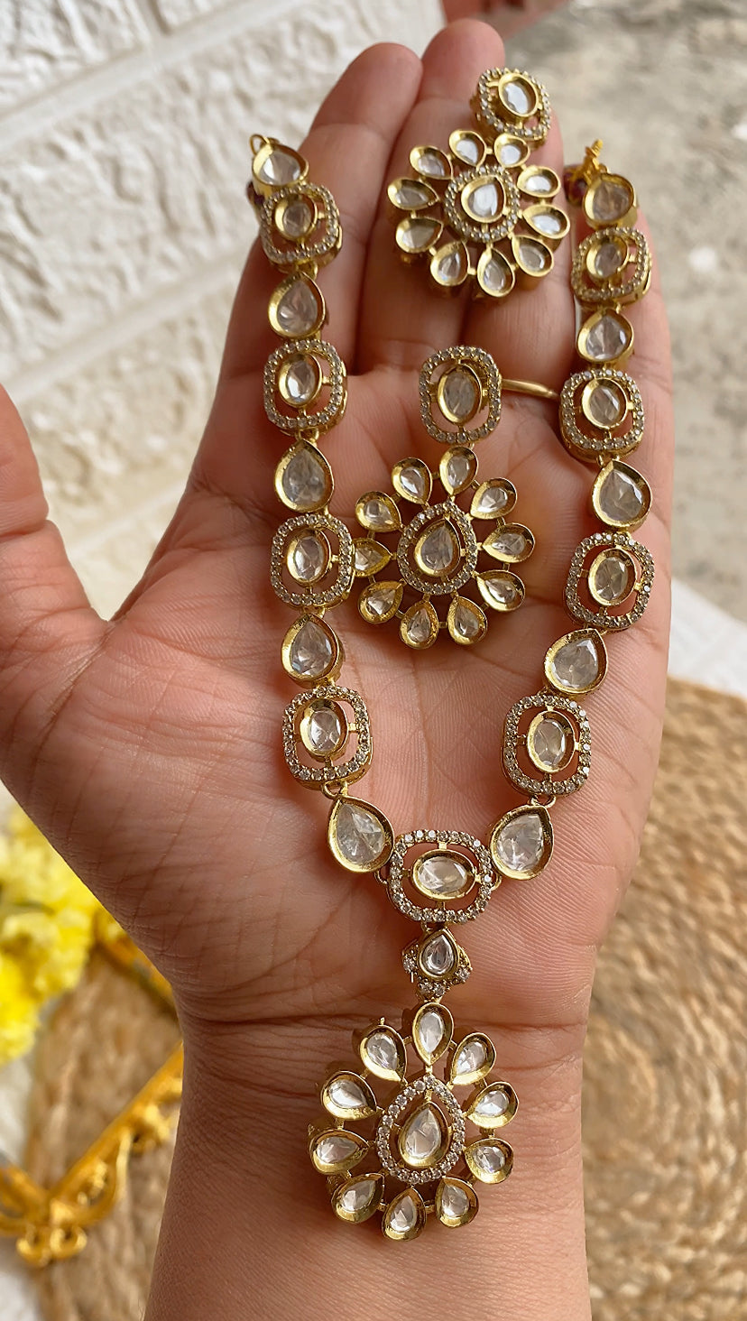 Gold jewelry set including necklace and earrings held in a hand against a textured beige background.